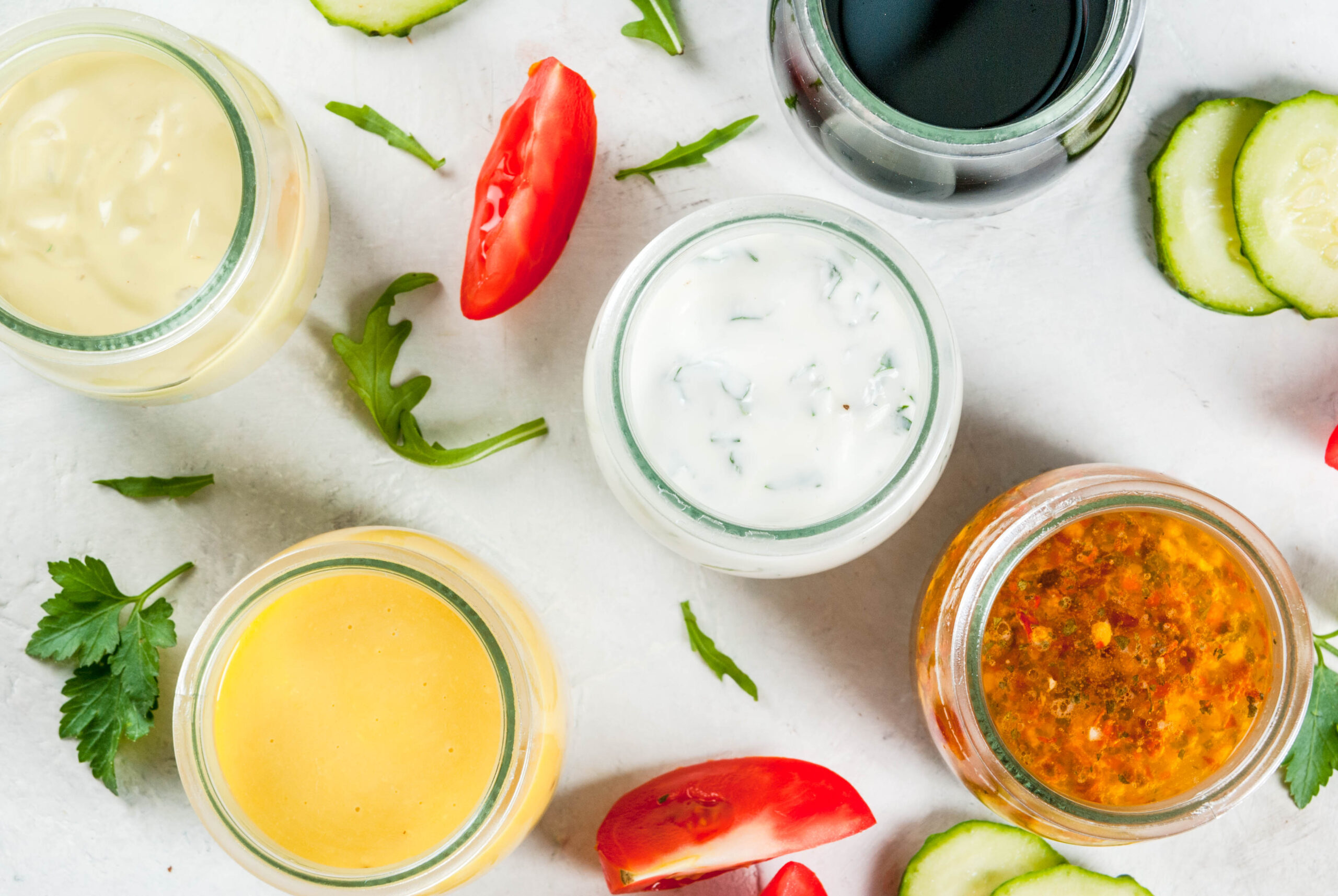 Set of dressings for salad: sauce vinaigrette, mustard, mayonnaise or ranch, balsamic or soy, basil with yogurt. Dark white concrete table, with greenery, vegetables for salad. Copy space top view